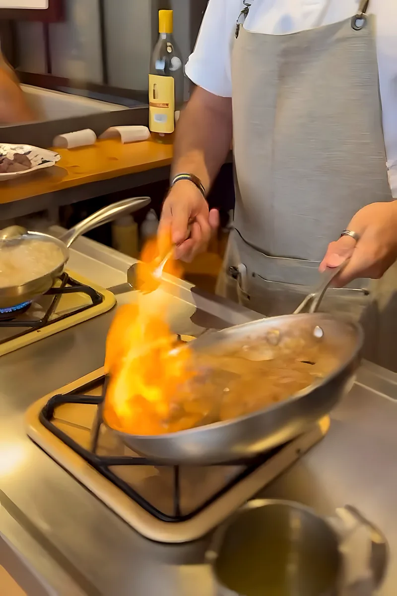 La llama sube en la sartén mientras el cocinero flambea, en el pase abierto a la sala